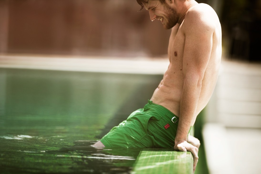 Man wearing green swimming trunks by Orlebar Brown for Monocle sitting at water's edge.