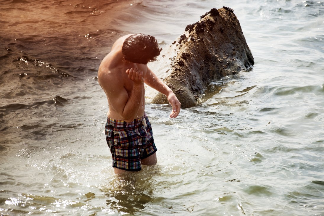 Man wading in the ocean wearing Polo Ralph Lauren swimming trunks near a large moss-covered rock.