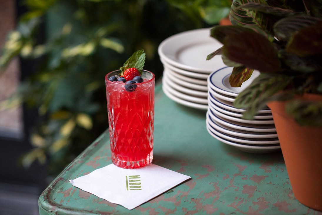 Red cocktail garnished with berries and mint leaf on table with stacked plates at Mardi restaurant.