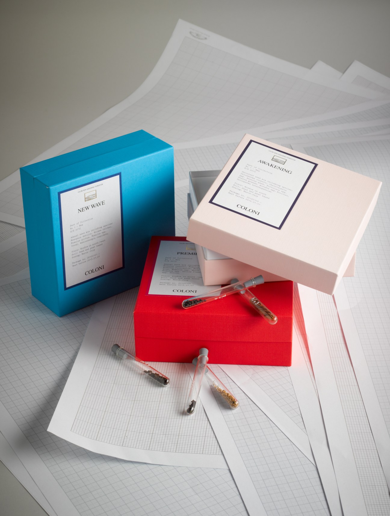 Coloni New Gardening seed collection boxes in blue, red and pink with labeled packets and glass seed vials on white grid paper.
