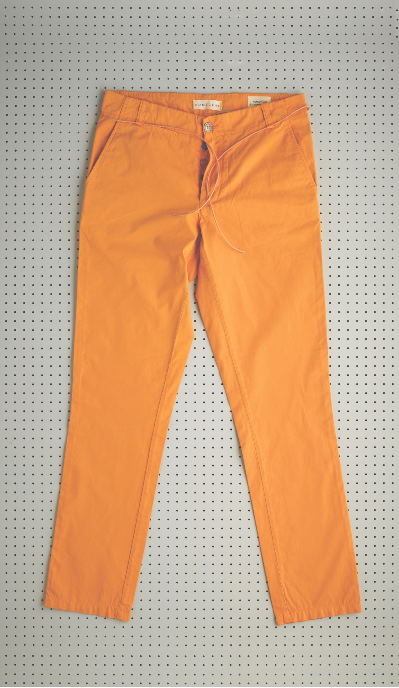 Bright orange chinos displayed flat against a perforated grey background.