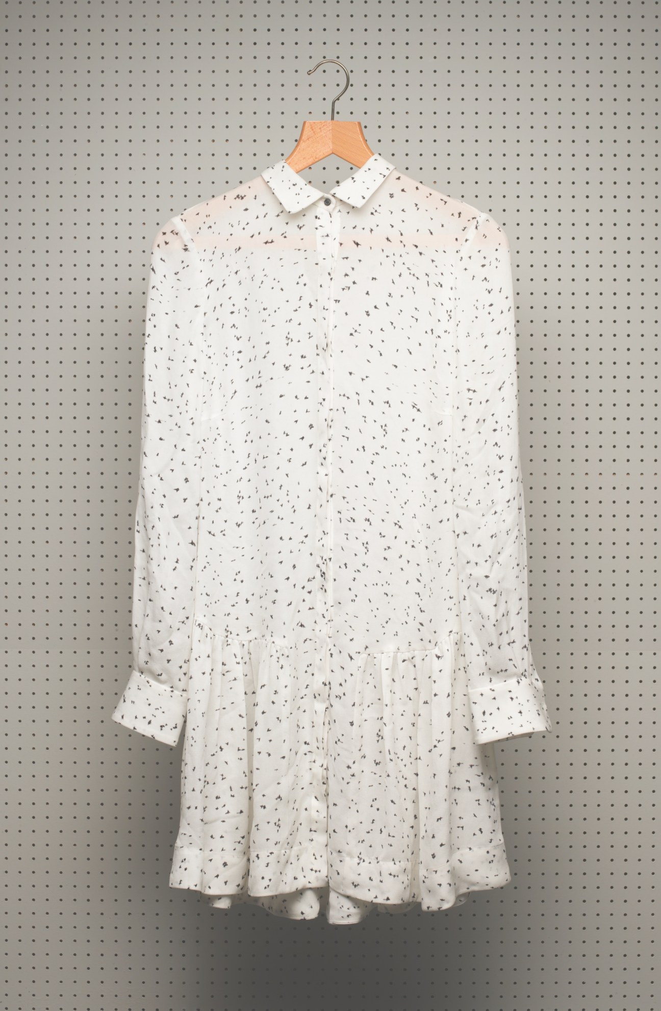 White silk dress with bird print and white collar on wooden hanger against gray pegboard.