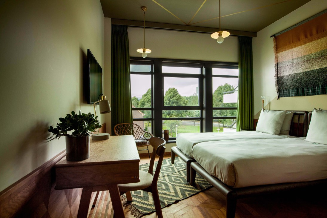 A modern hotel bedroom with pale green walls, large windows overlooking greenery, and mid-century furniture including wooden desk and chairs.