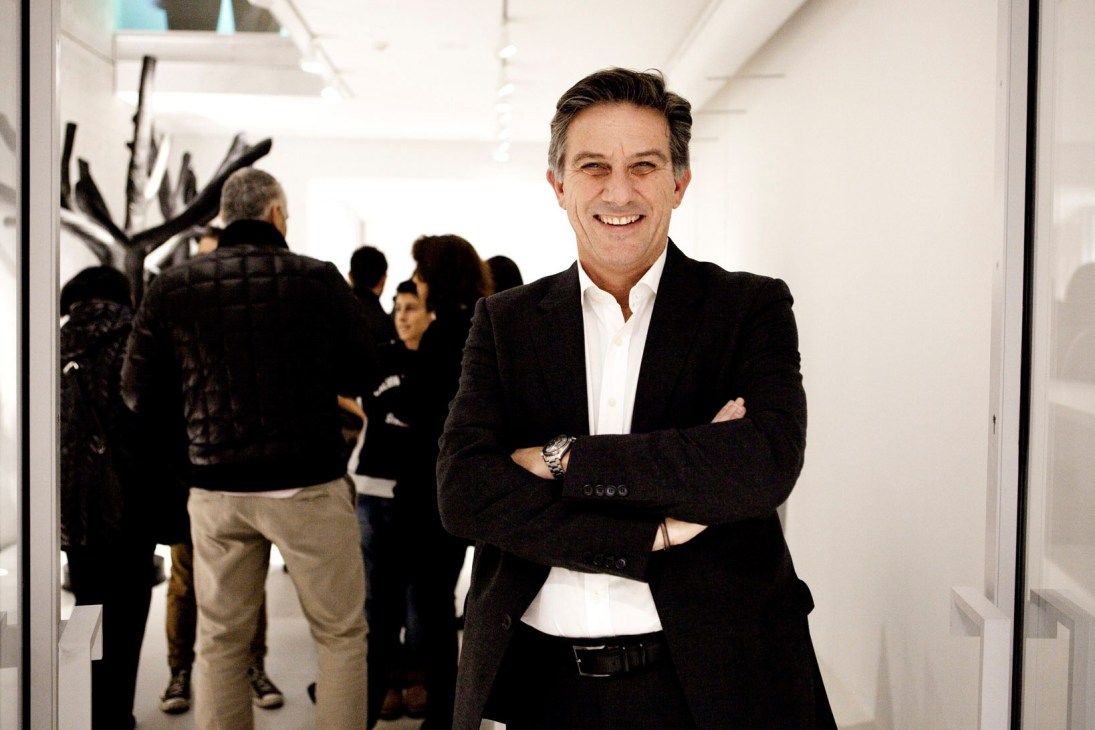 Man in black blazer and white shirt smiling at camera in modern white gallery space with visitors in background.