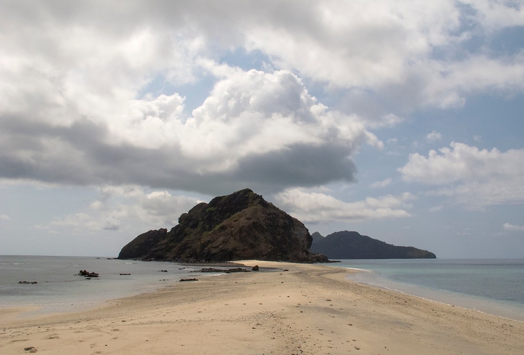 A sandy beach stretches toward a rocky island rising from the sea under a cloudy sky.