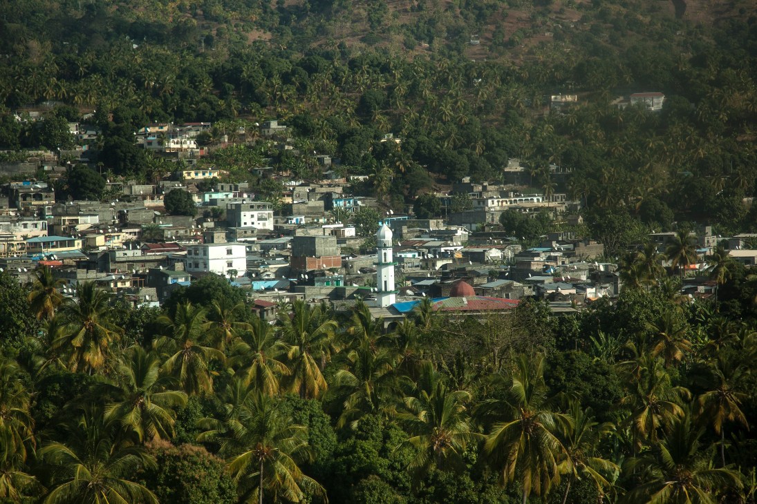Aerial view of a hinterland town on Anjouan with a white minaret rising above densely packed buildings surrounded by coconut palms and tropical vegetation.