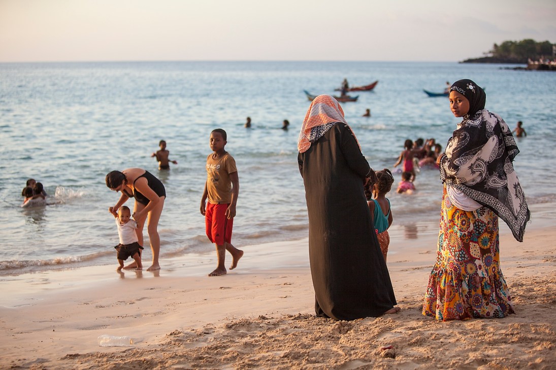 Women and children on a sandy beach in Maroni, Grand Comoros, with fishing boats visible offshore.