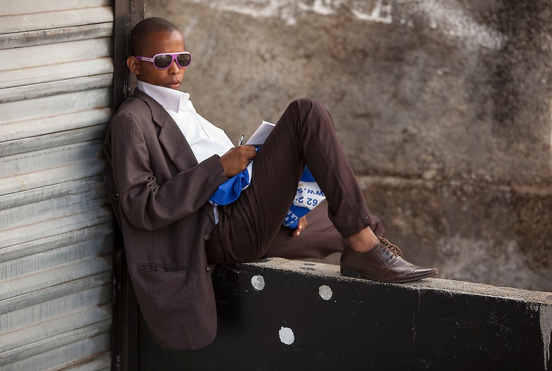 Man in brown suit and pink sunglasses sitting against weathered wall holding passport.