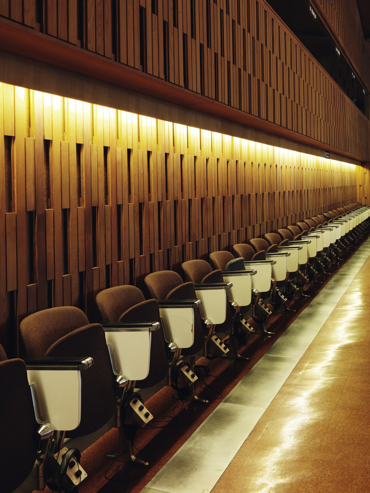 Rows of brown bucket seats with cream-colored side tables line a modernist conference hall with wooden slat walls and ambient lighting.
