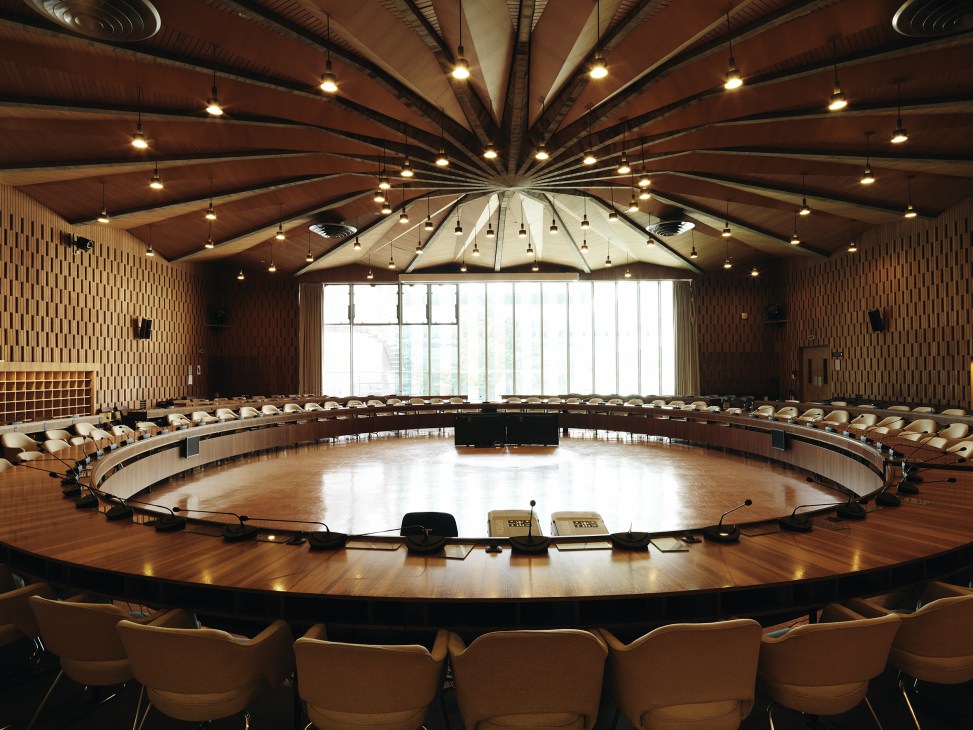 Modernist conference room with circular wooden table, radiating ceiling beams, and bucket seats arranged in a semicircle.