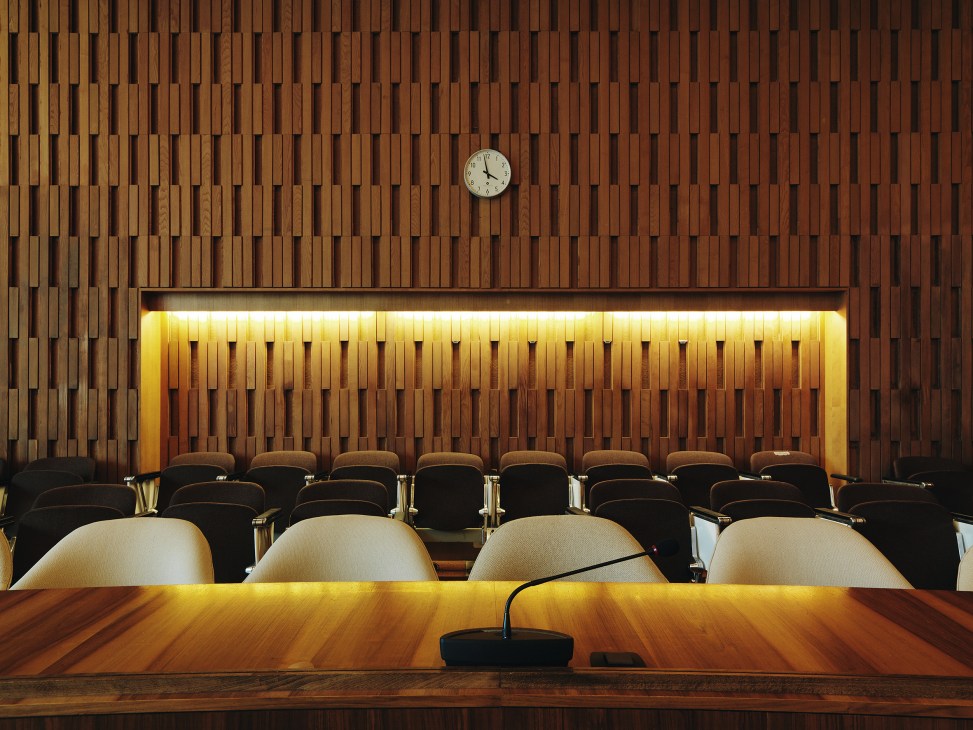 Modernist conference hall with wooden slat wall paneling, cream-colored bucket seats, and a wall clock above an illuminated alcove.