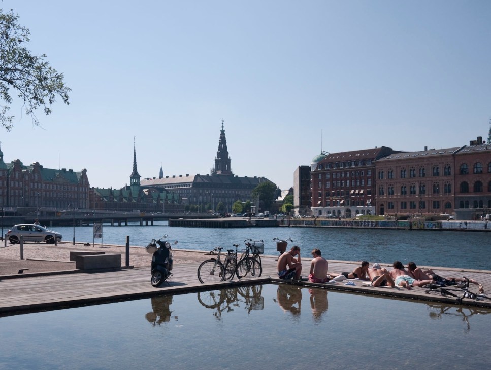 Copenhagen waterfront with people relaxing on a wooden pier, bicycles, historic buildings and church spires across the water.