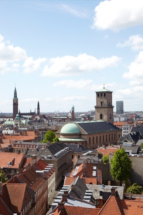 Aerial view of Copenhagen showing historic red-roofed buildings and church spires under blue sky.