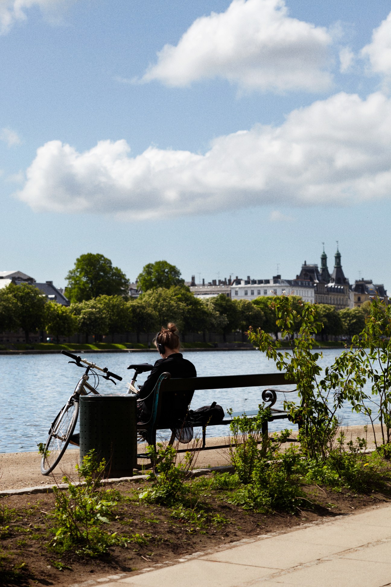 Person sitting on a bench with a bicycle by a riverside in Copenhagen, with historic buildings across the water.