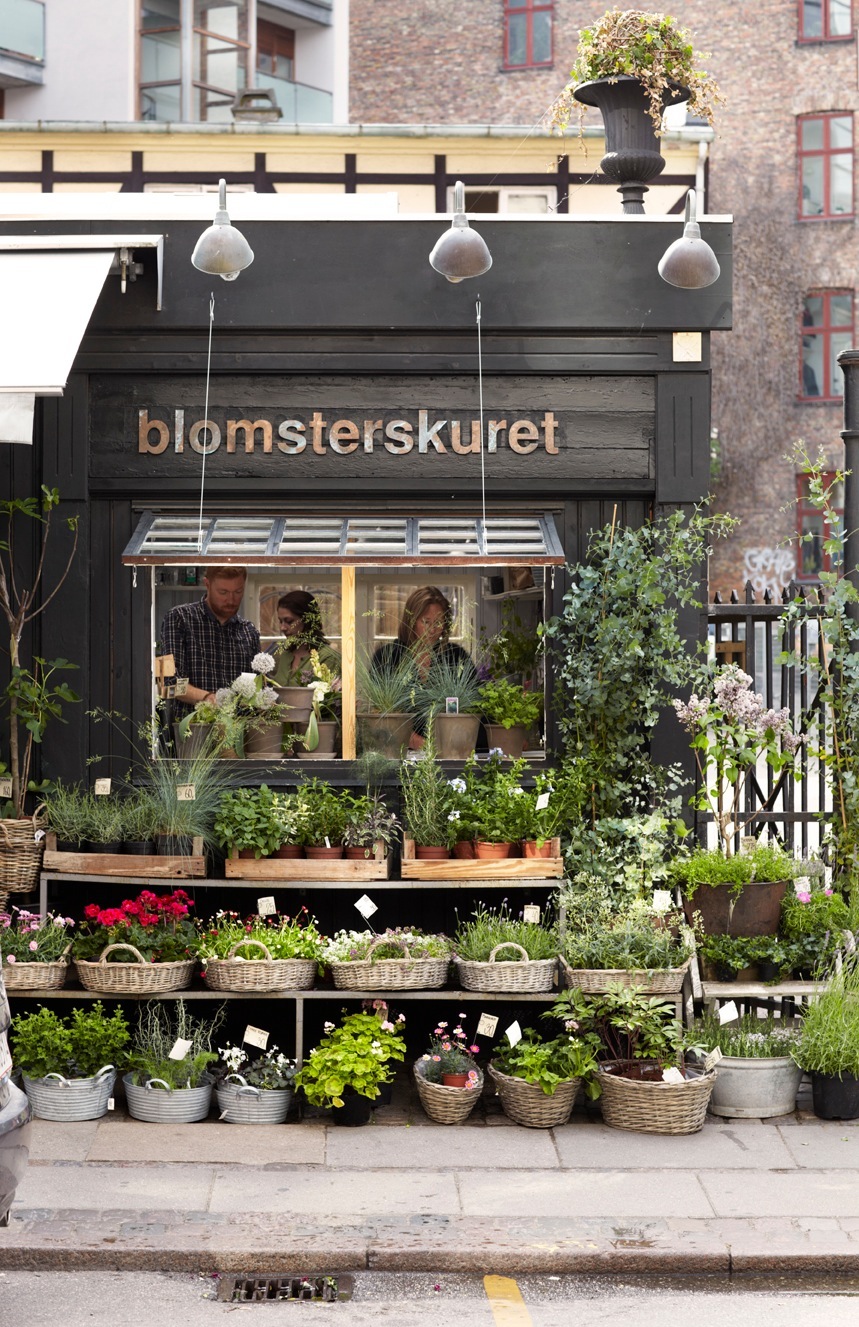 Blomsterskuret flower shop storefront with black facade, plants displayed outside, and brick building behind.