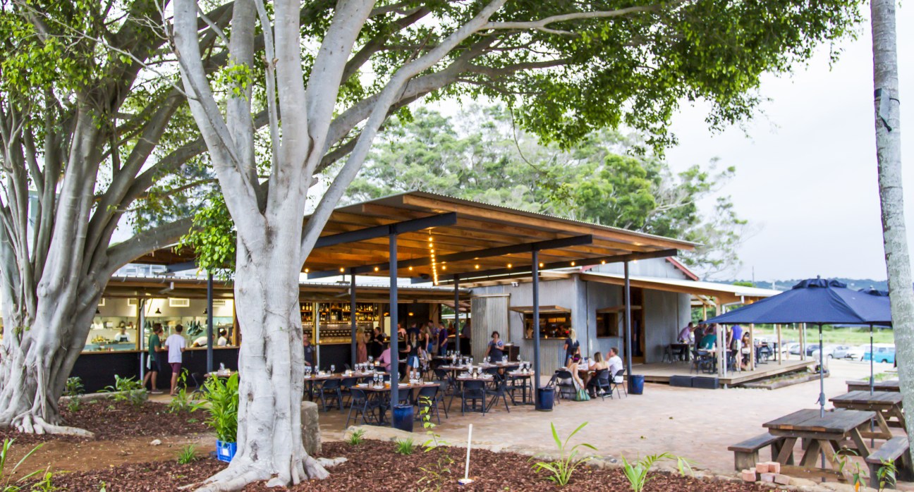 The Farm restaurant in Byron Bay with large white gum trees, covered dining areas, and visitors seated at tables under wooden structures.