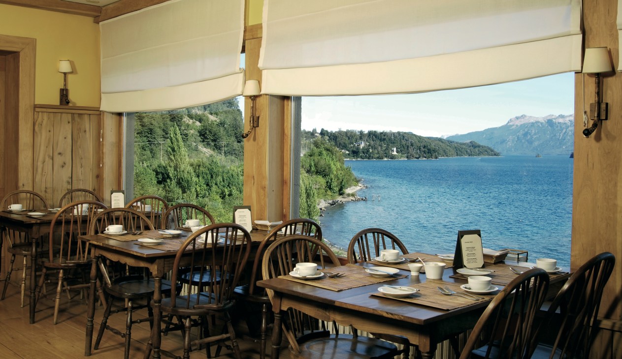 Dining room with wooden tables and chairs overlooking Lake Nahuel Huapi and snow-capped Andes mountains through large windows.