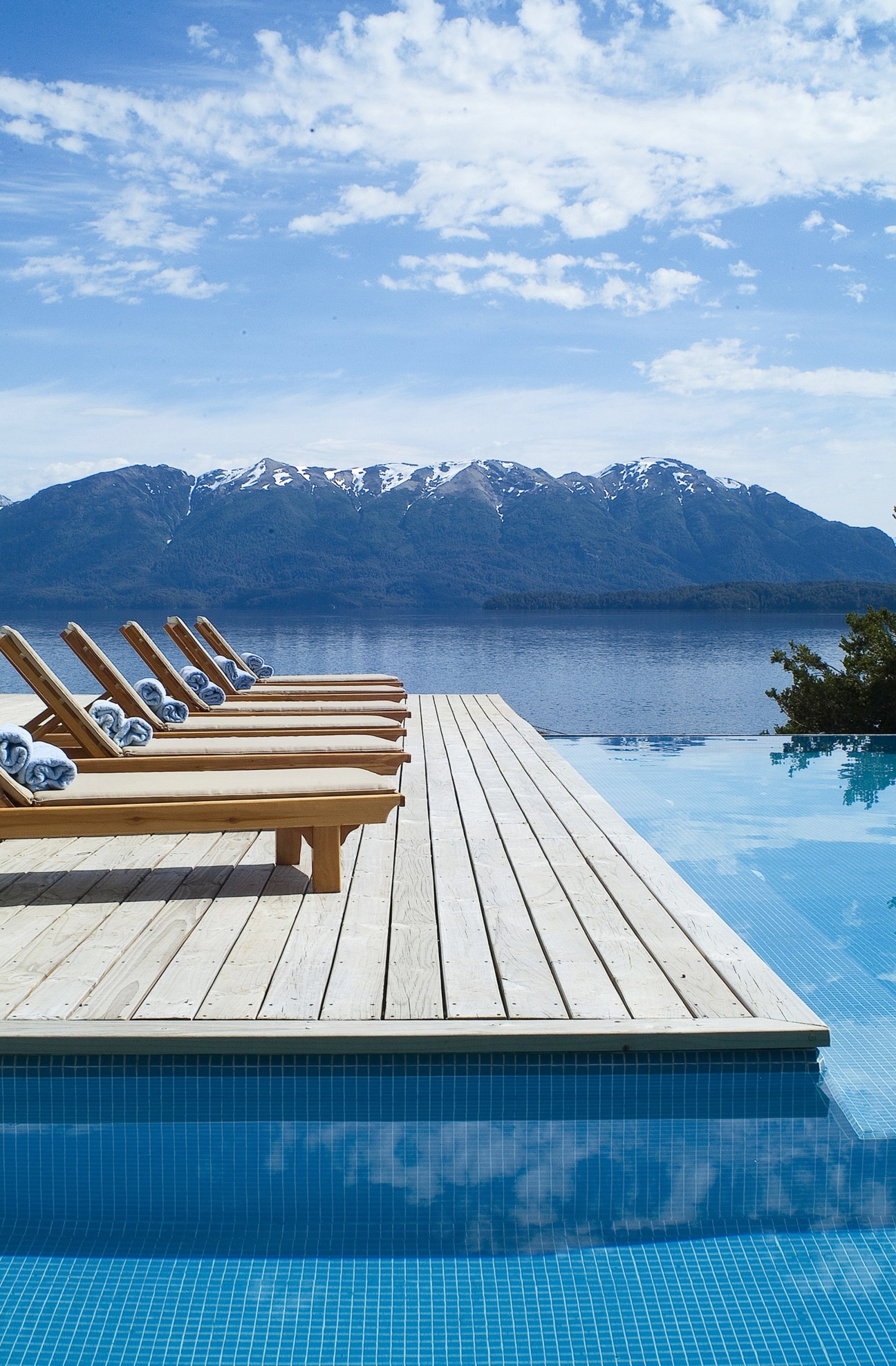 Wooden lounge chairs on a deck overlooking an infinity pool, lake, and snow-capped mountains in Patagonia.