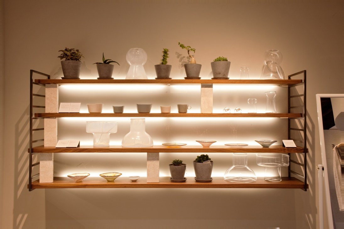 Illuminated wooden shelving displaying Three cosmetics products, glass vessels, potted plants and ceramic containers.
