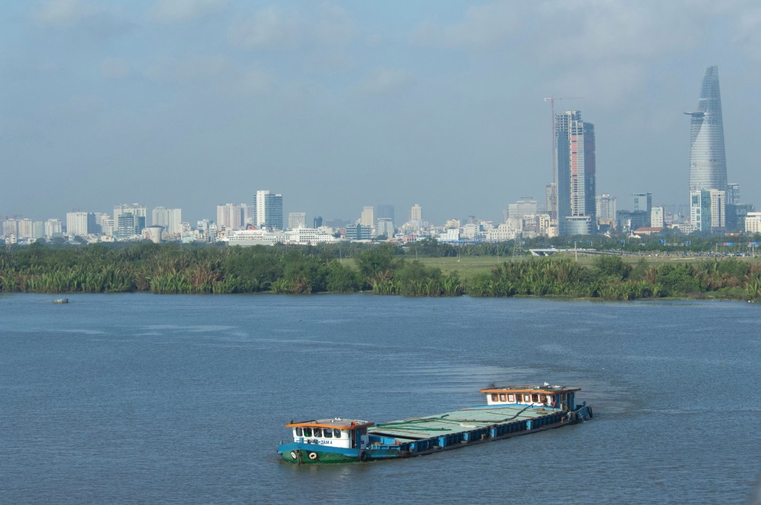 Ho Chi Minh City skyline with modern towers across the Saigon River and a traditional wooden cargo boat in foreground.