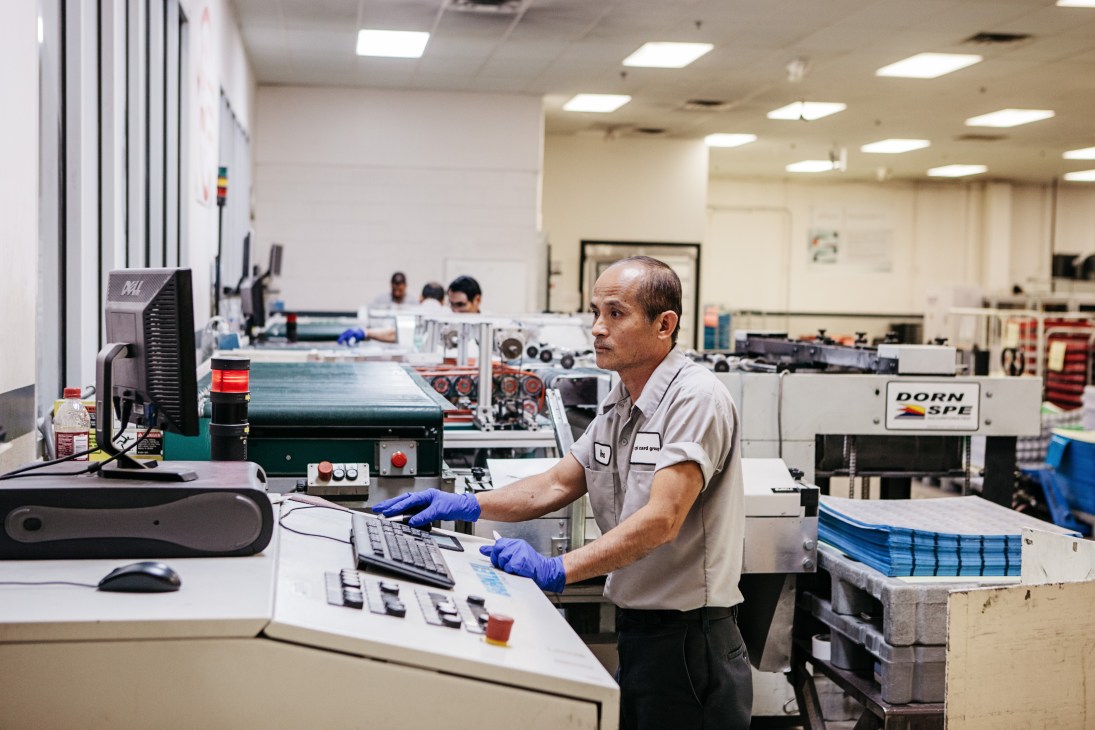 Worker in blue gloves operating laminating machinery at CPI Card Group manufacturing facility.