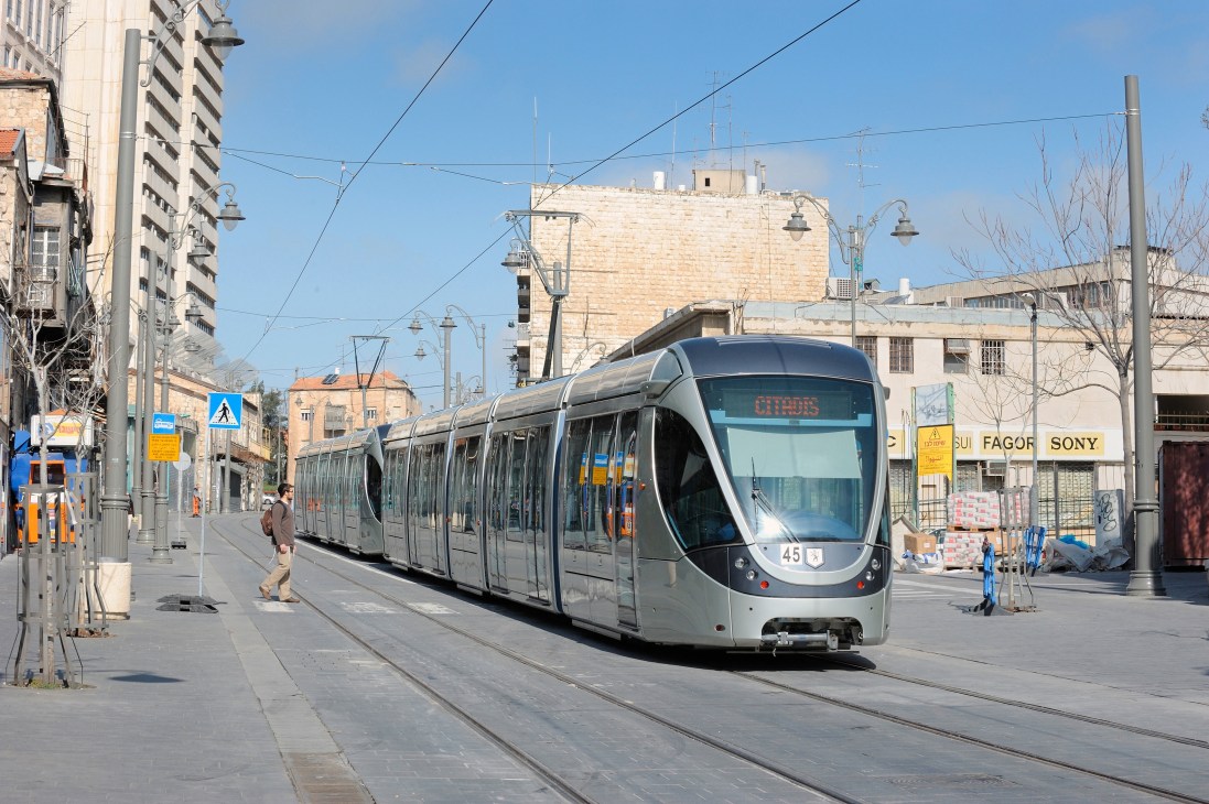 Modern light rail tram traveling through Jerusalem's streets with historic stone buildings and clear blue sky in background.
