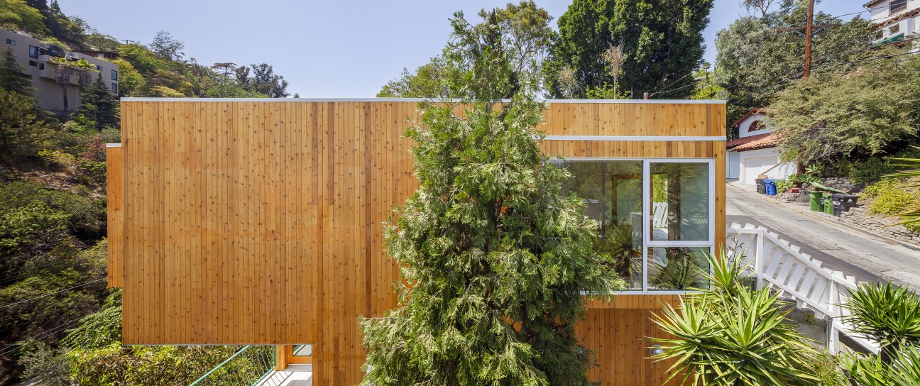 Cedar-clad compact house with large windows on a hillside surrounded by vegetation.