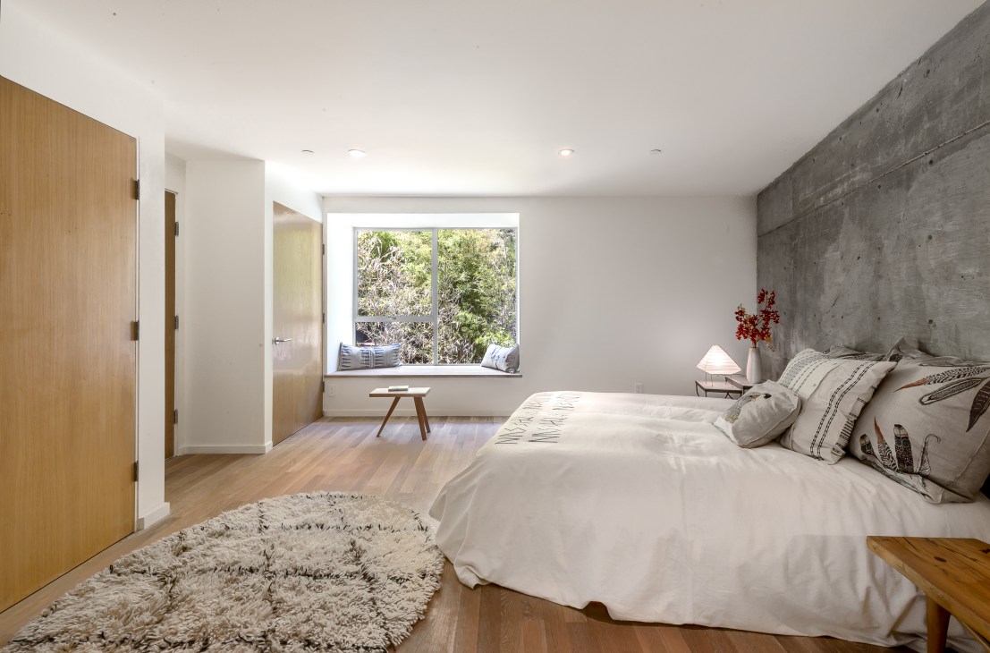 Bright bedroom with white oak flooring, exposed concrete wall, and bay window with built-in seating overlooking landscaped slope.