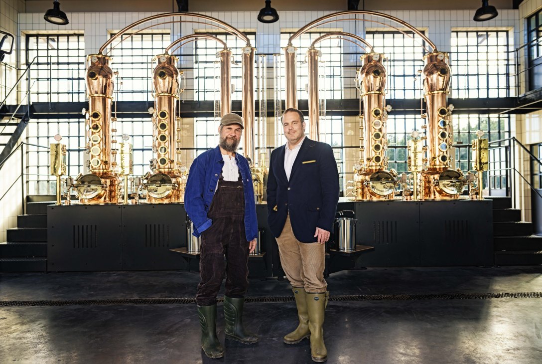 Monkey 47 distiller Christoph Keller and founder Alexander Stein stand before large copper distilling equipment.