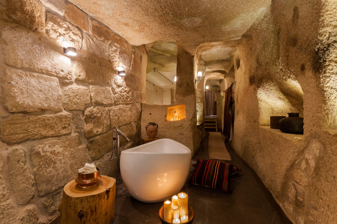 Ancient cave bathroom with white soaking tub, stone walls, and candlelit ambiance.