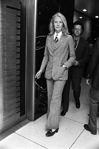 Black and white photograph of a woman in a masculine tailored pantsuit walking through a corridor with a man behind her.