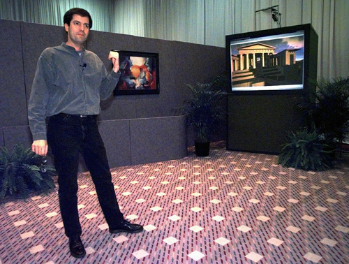 Man in casual business attire presenting in an office setting with display screens.