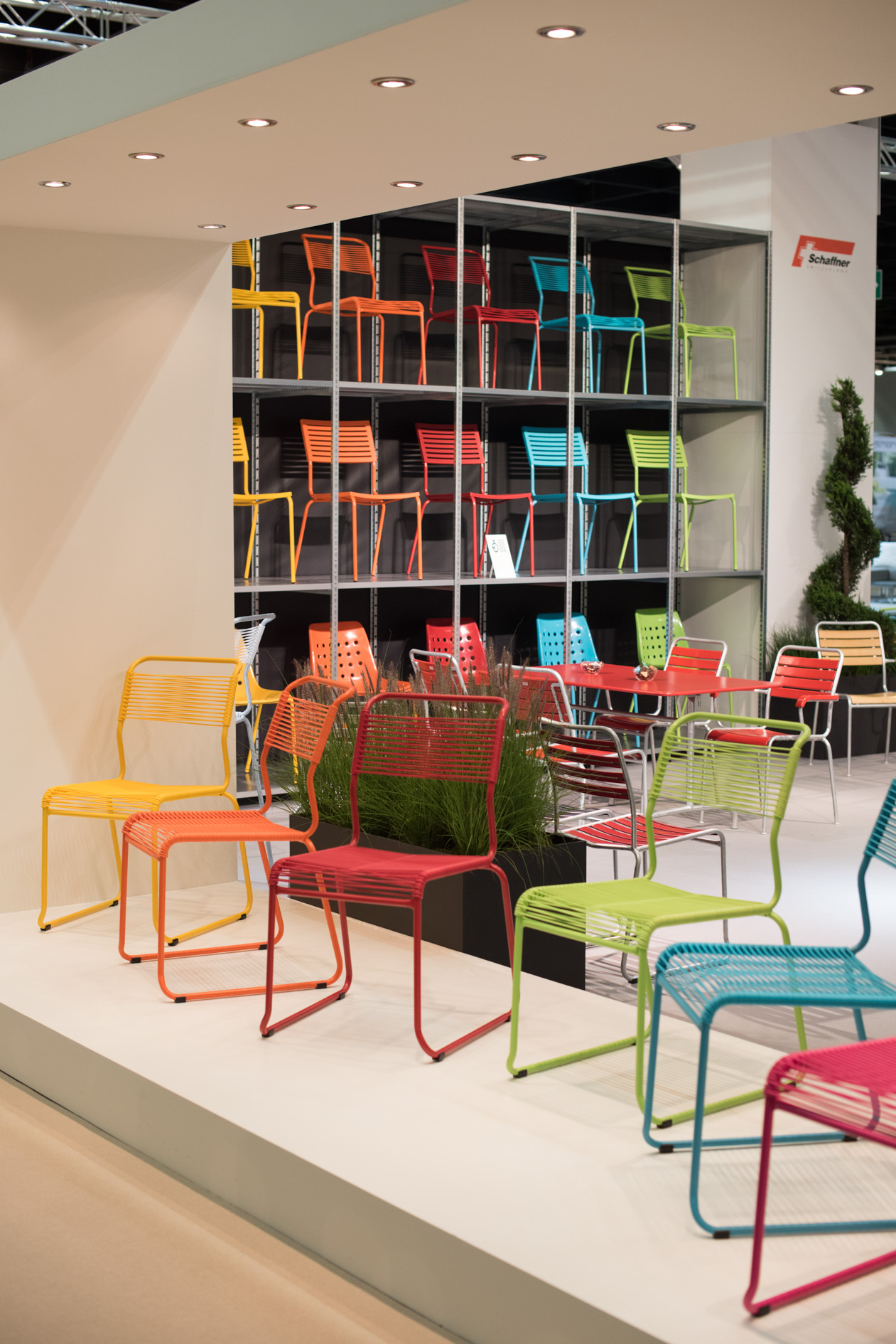 Schaffner's colorful metal Spaghetti chairs displayed at a trade fair booth with shelving unit behind.