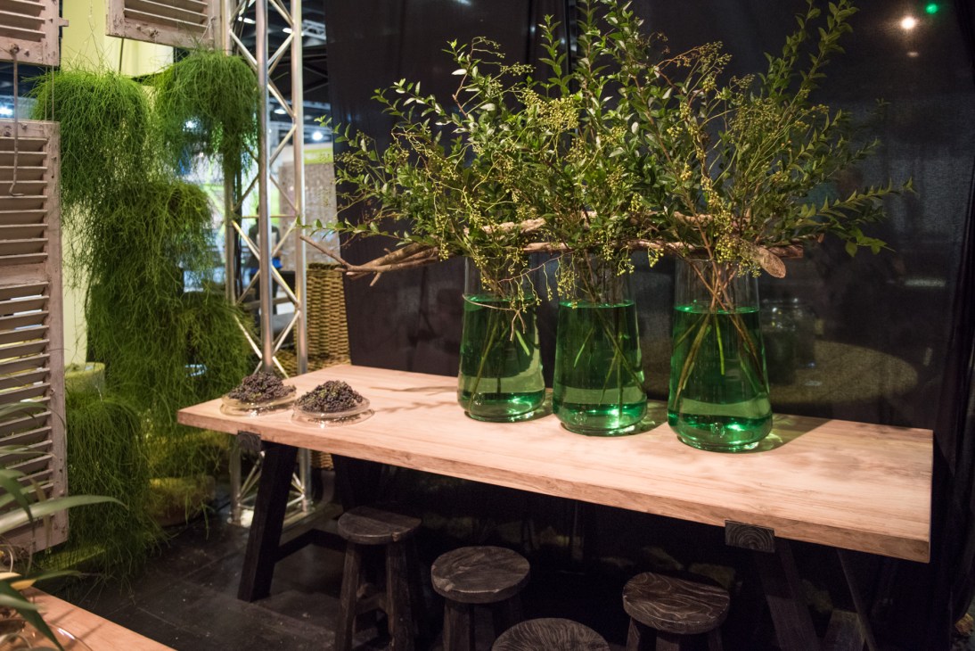 Gommaire's wooden display table with green glass vases holding branches and scattered seeds.