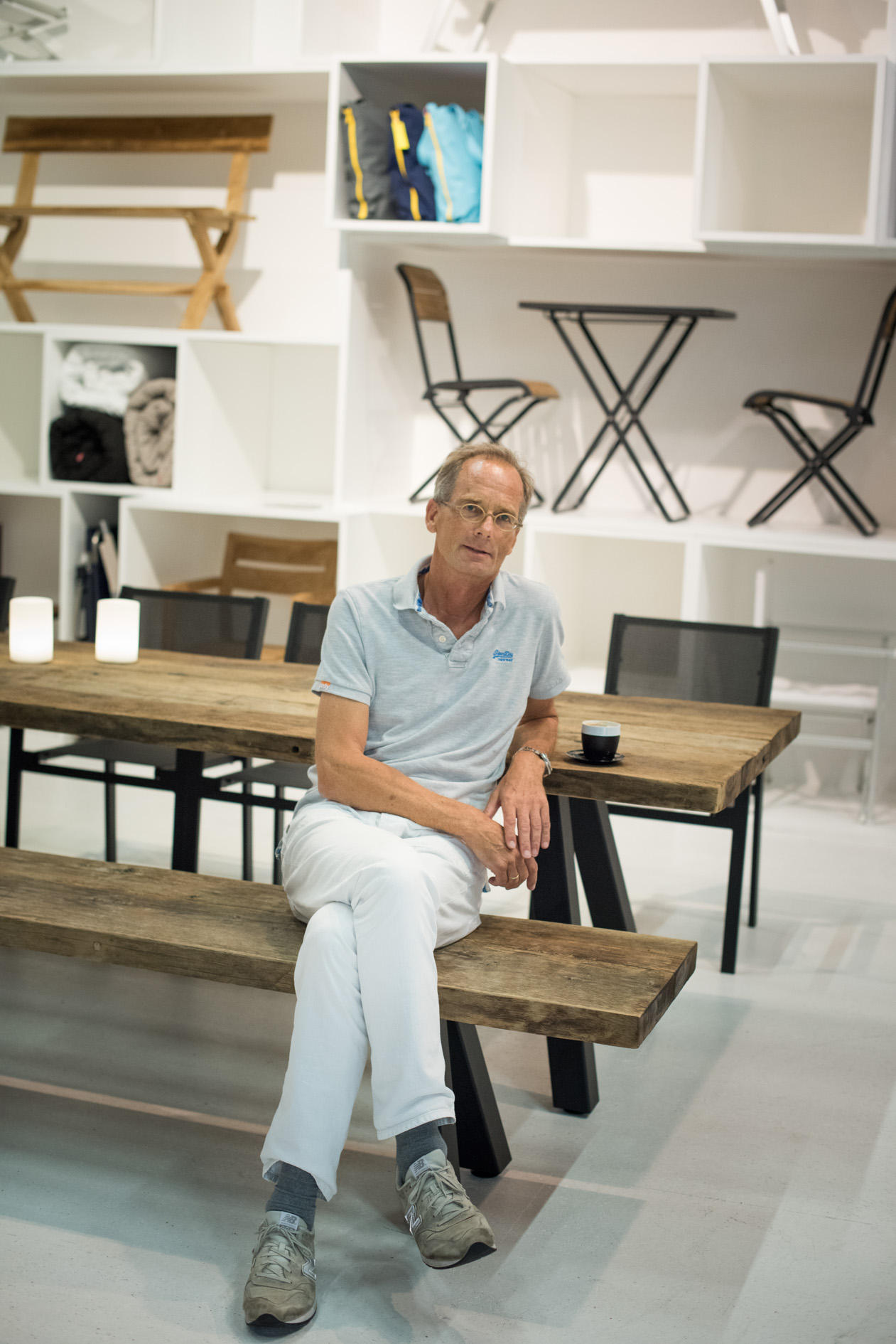 Jan Kurtz sits on a wooden bench at his furniture booth displaying chairs and shelving.