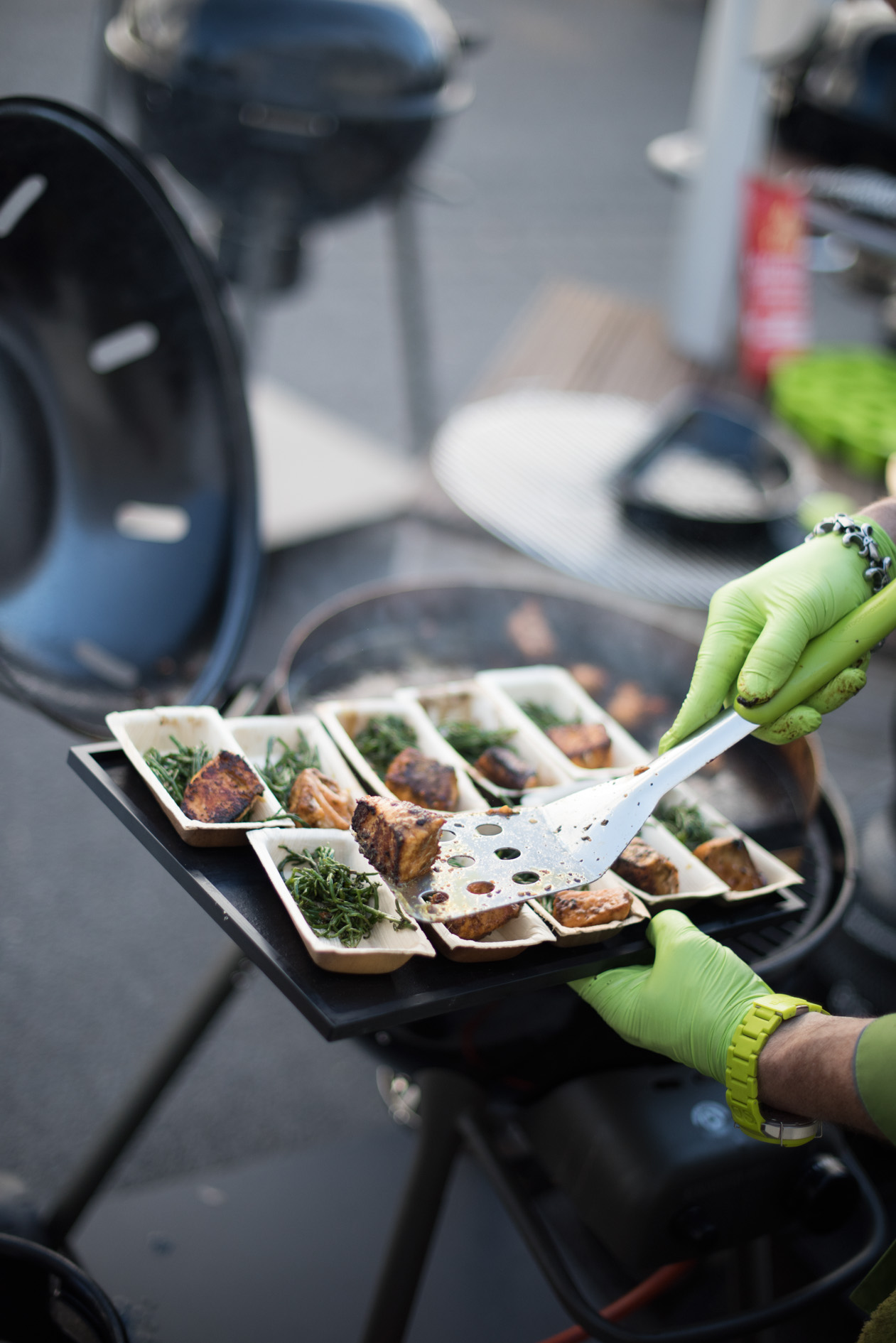 Person in bright green gloves serving grilled meat samples on white platters at the garden fair.
