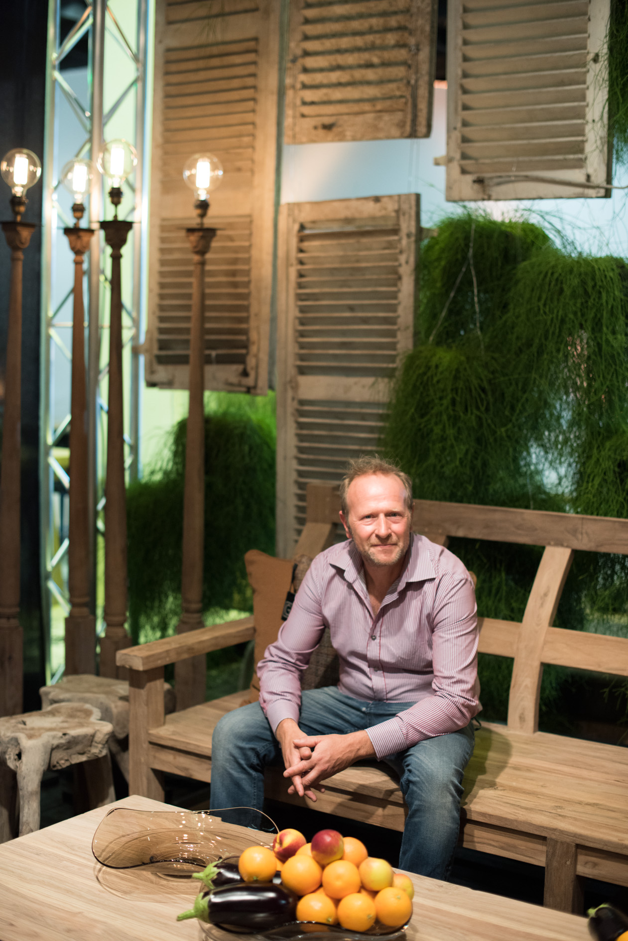 Designer Gommaire Cleybergh sits on a wooden chair at a trade fair display with vintage shutters and plants.