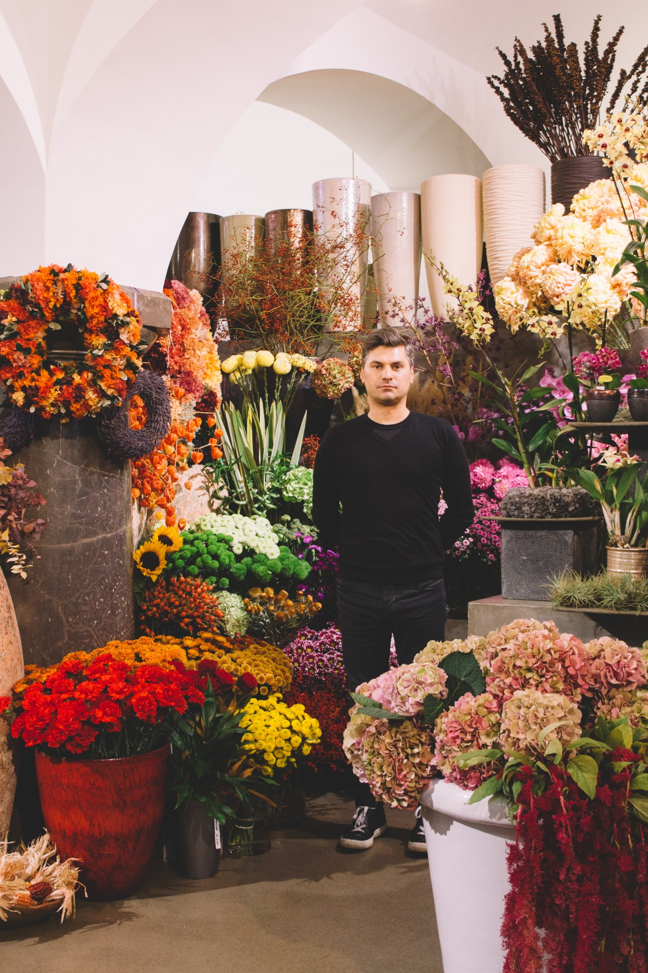 A man in a black sweater stands surrounded by vibrant flowers at Cvetlicarna Emporia florist shop in Ljubljana.