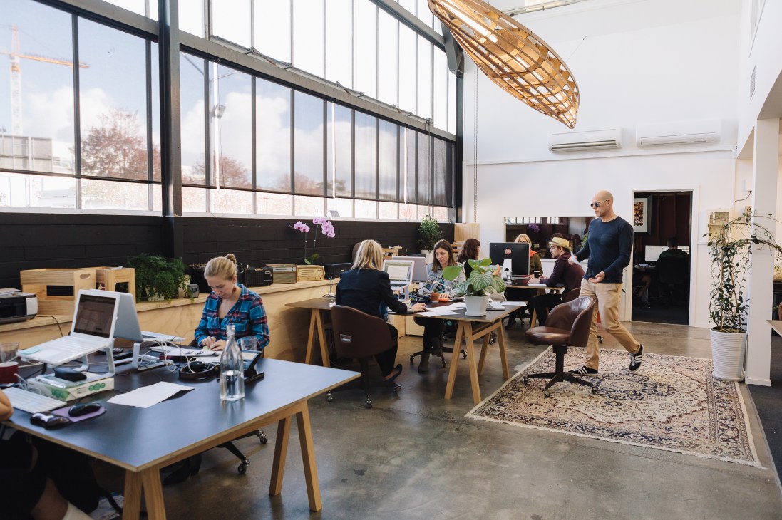 Open-plan creative studio with workers at desks, large windows, wooden furniture, and industrial ceiling in renovated workshop space.