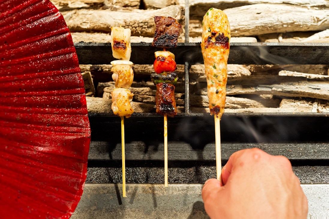 Grilled chicken skewers cooking on a charcoal grill with a red fan visible on the left.