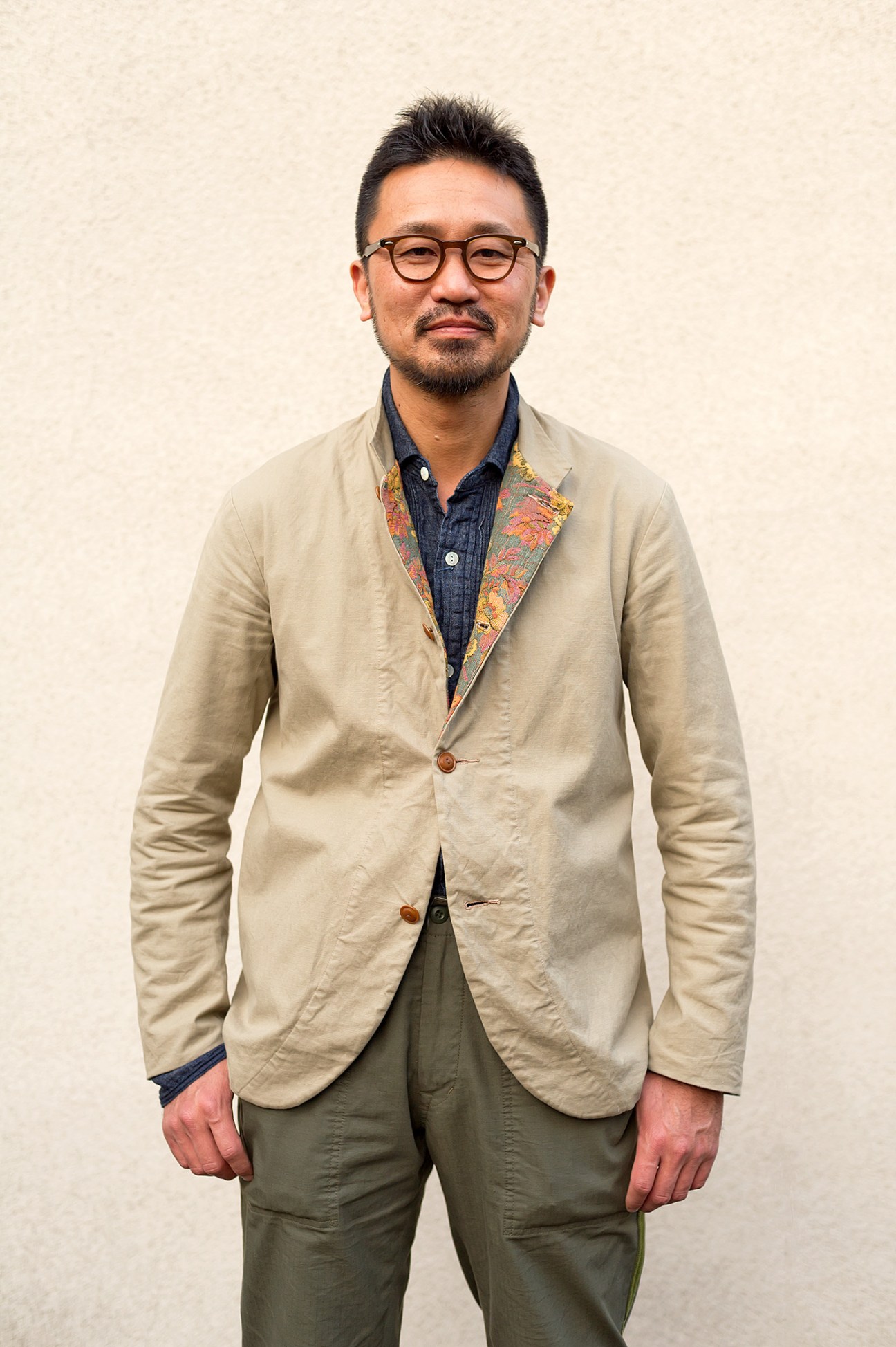 Kenichi Kusano, Baracuta's designer, stands in a neutral pose wearing a beige blazer with a patterned collar and glasses.