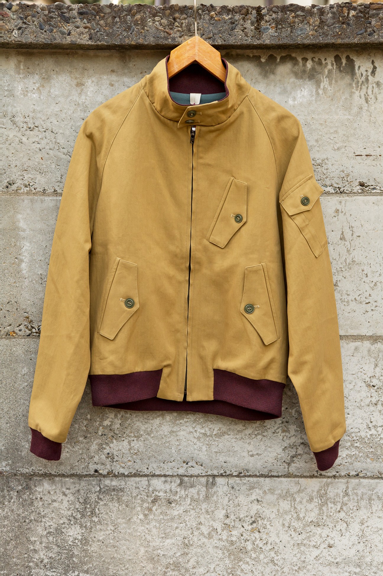 Mustard-yellow Baracuta G9 jacket with burgundy ribbed cuffs and collar on wooden hanger against concrete wall.