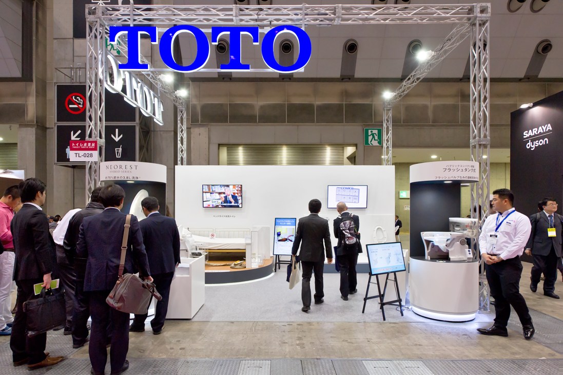 Toto's large exhibition booth at Toilet Tokyo convention with blue signage and visitors in business attire.