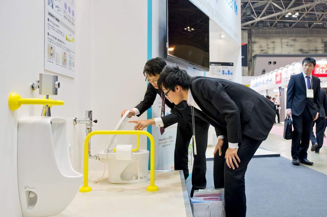 Two people examine a small white toilet with yellow safety rails at a trade show booth.