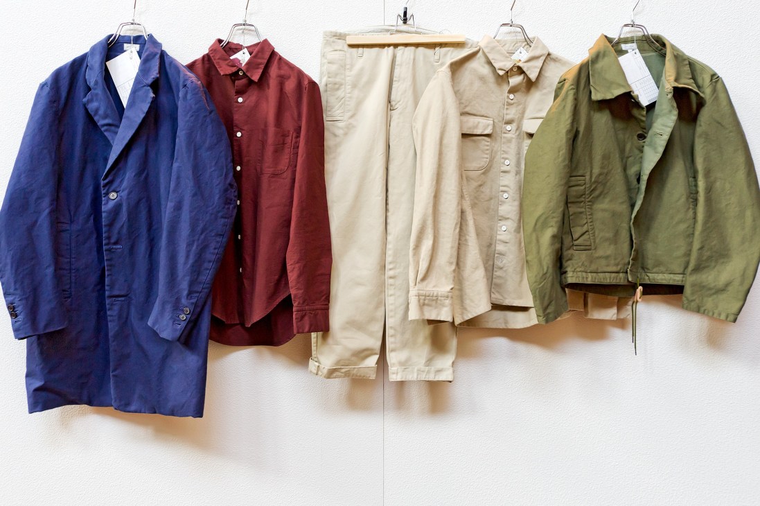 Five cotton basics garments in blue, burgundy, cream and olive displayed on hangers against a white wall.