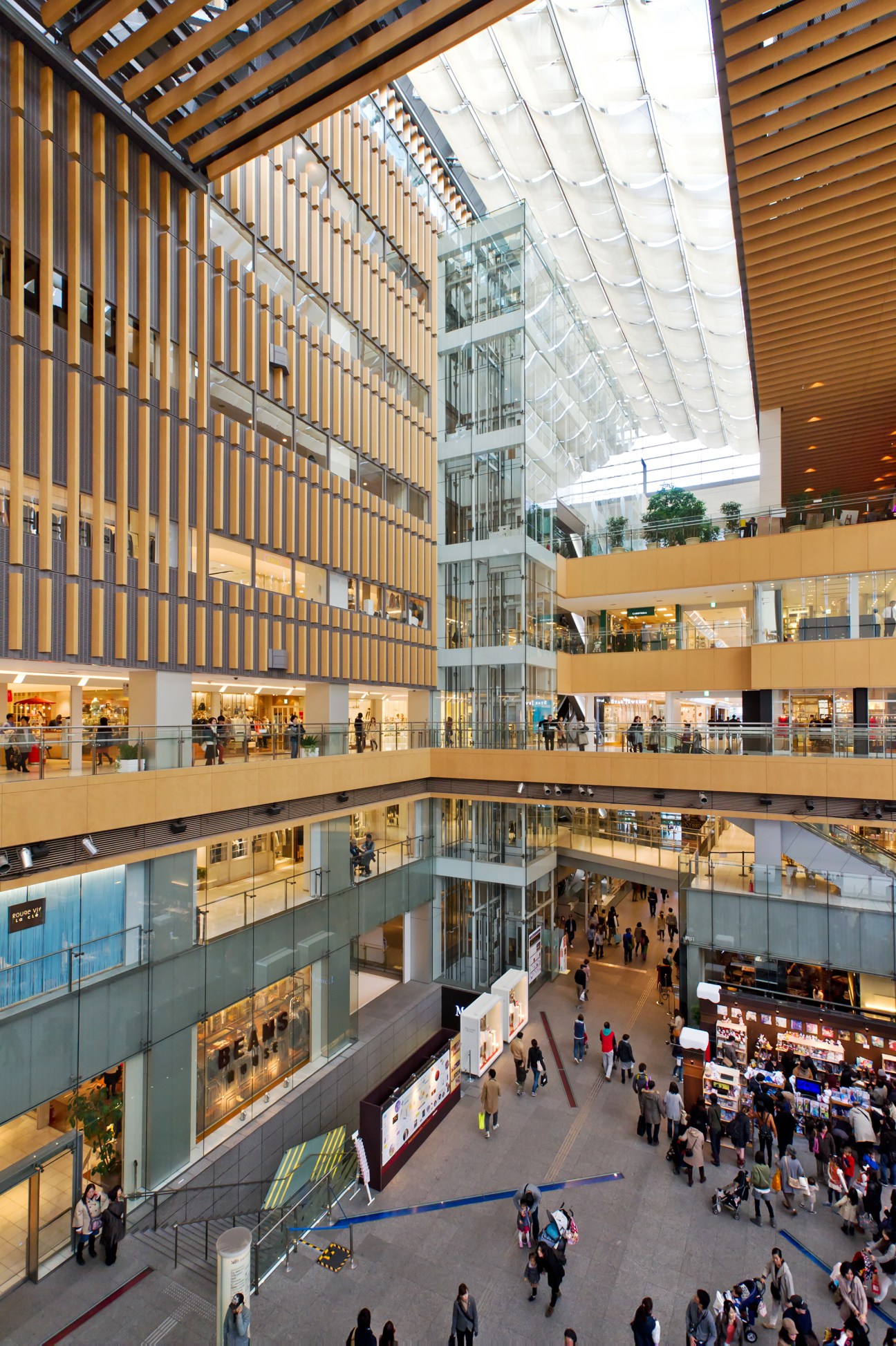 Interior of Marunouchi Building showing multiple retail levels with wooden lattice walls, glass railings, and shoppers across floors.