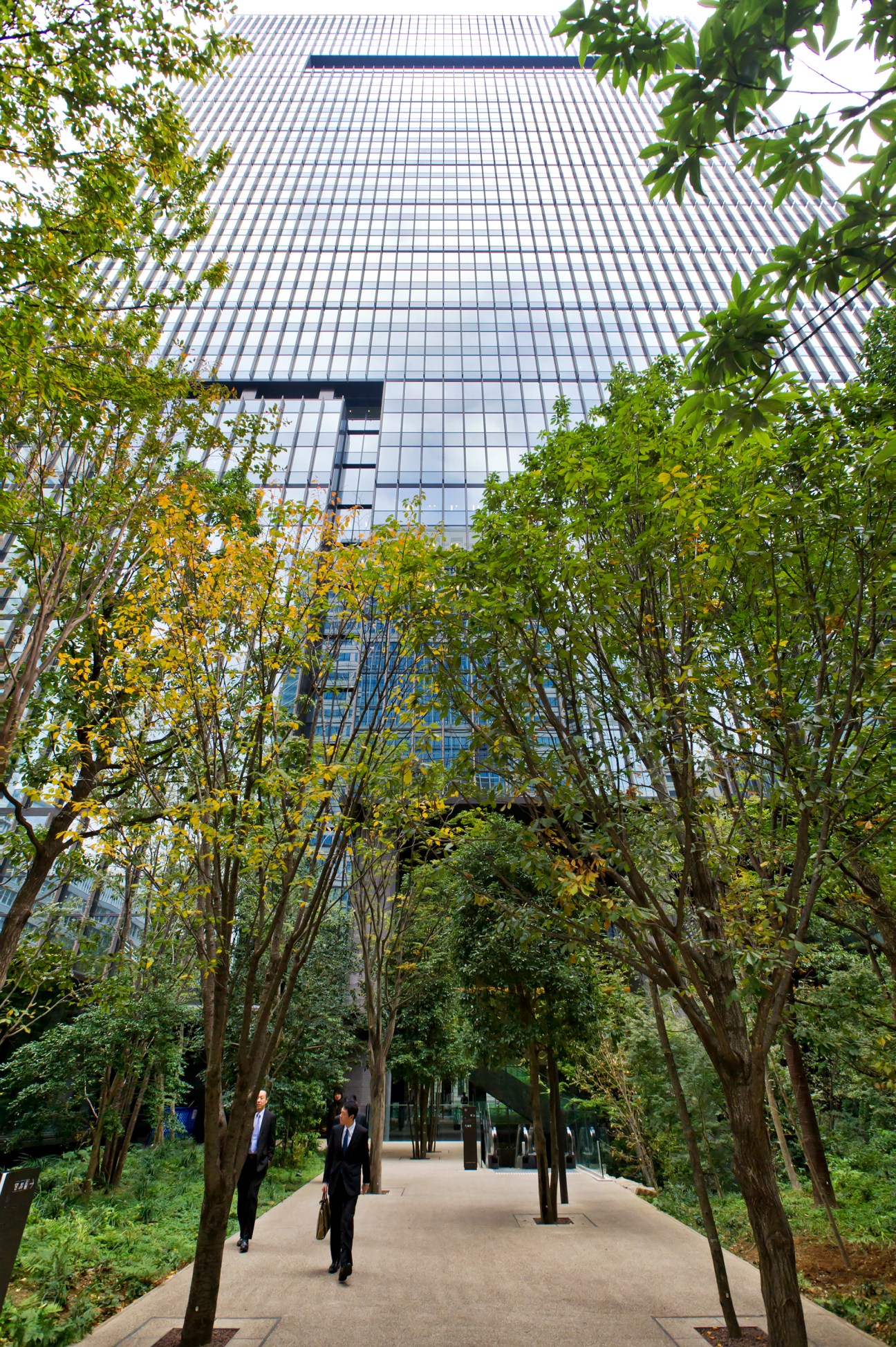 Otemachi Tower rises behind a tree-lined pedestrian walkway in Tokyo's Marunouchi district.