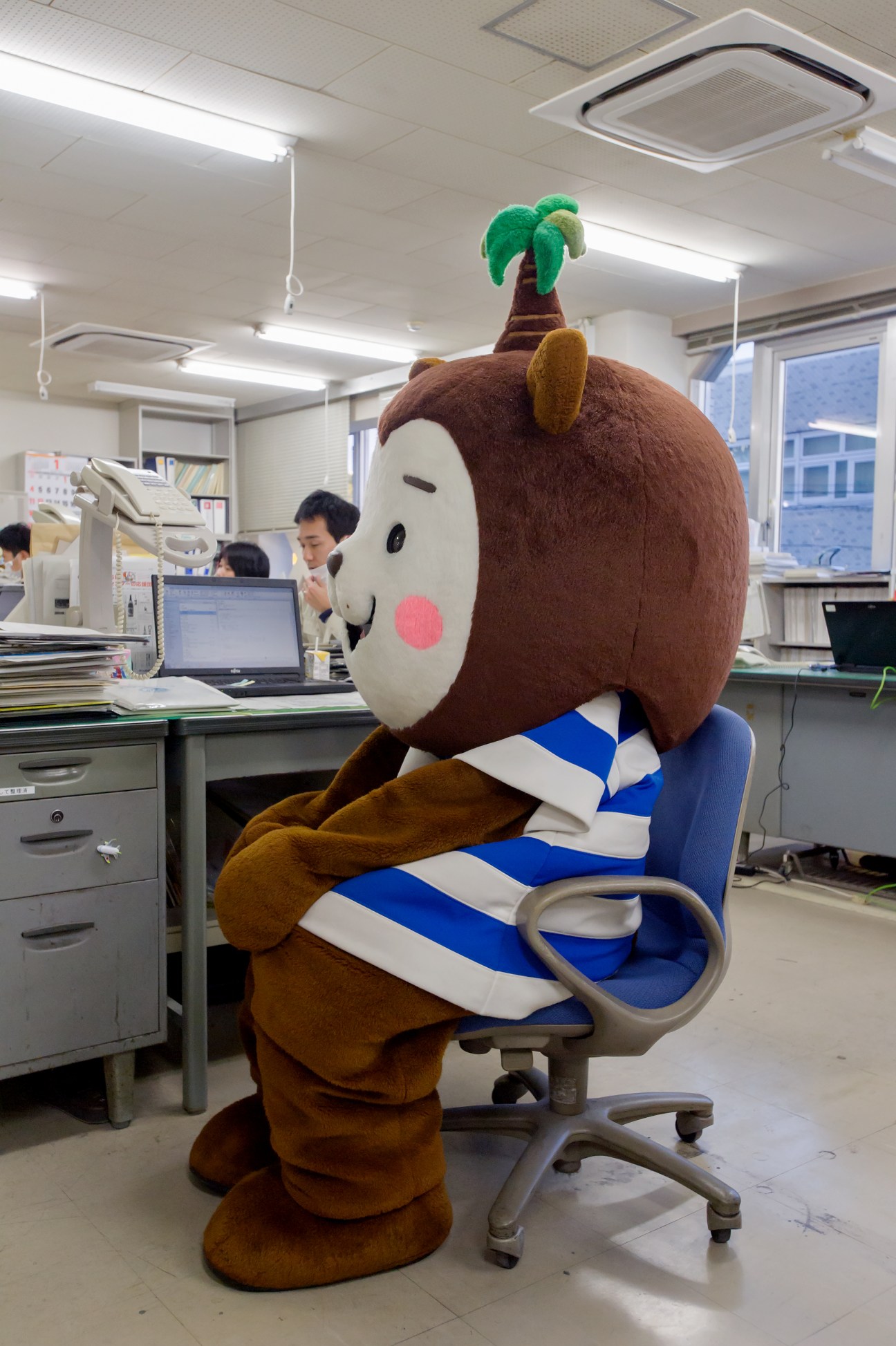 Muu-chan mascot character sits at desk in tourism office while staff work in background.