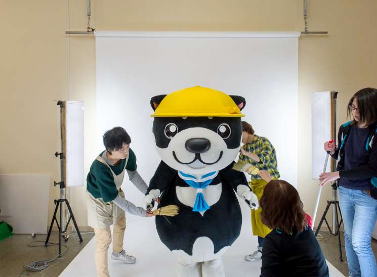 A black and white mascot character with a yellow hard hat poses for photographs with people around it in a studio setting.