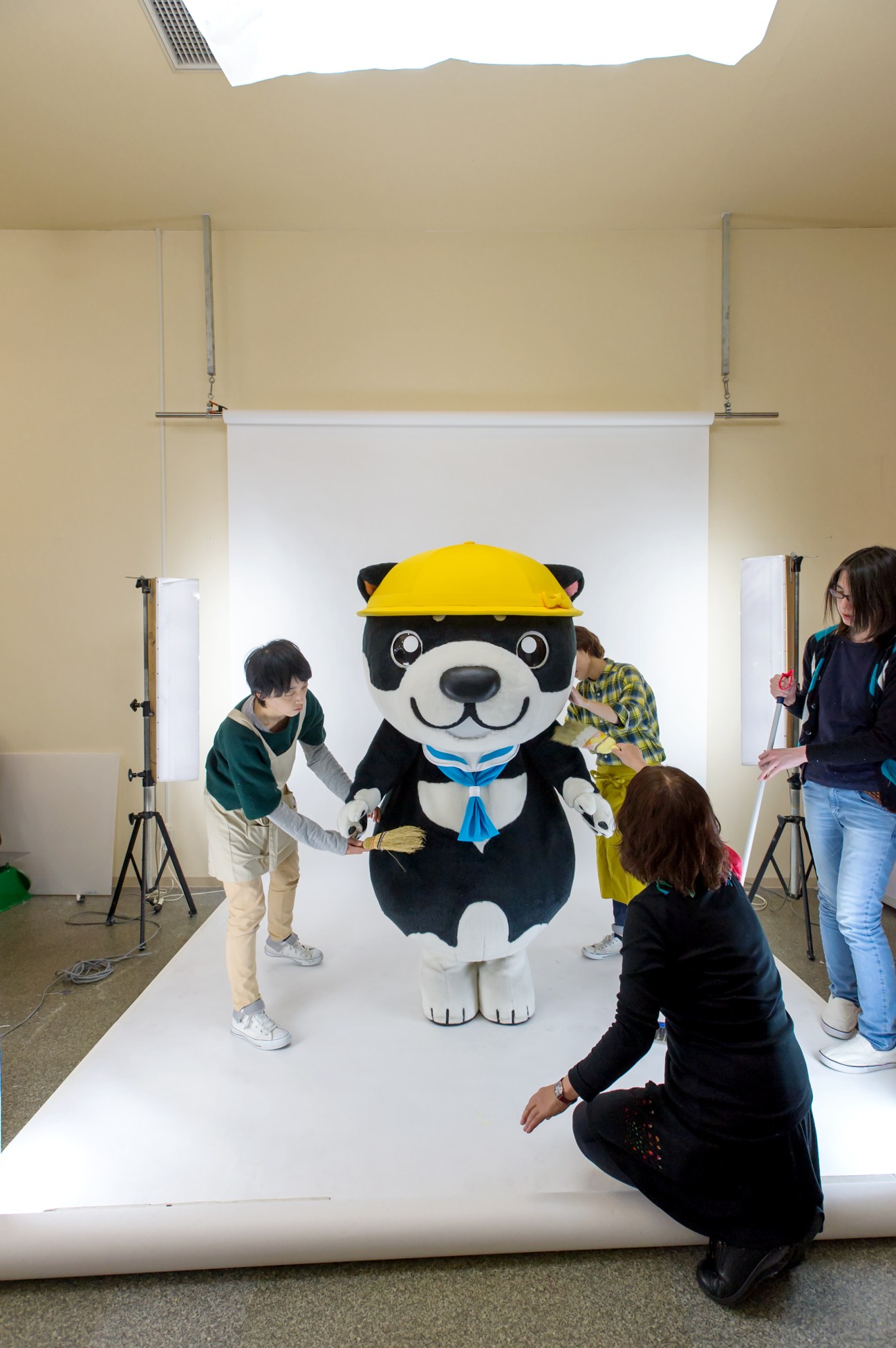 A black and white mascot character with a yellow hard hat poses for photographs with people around it in a studio setting.